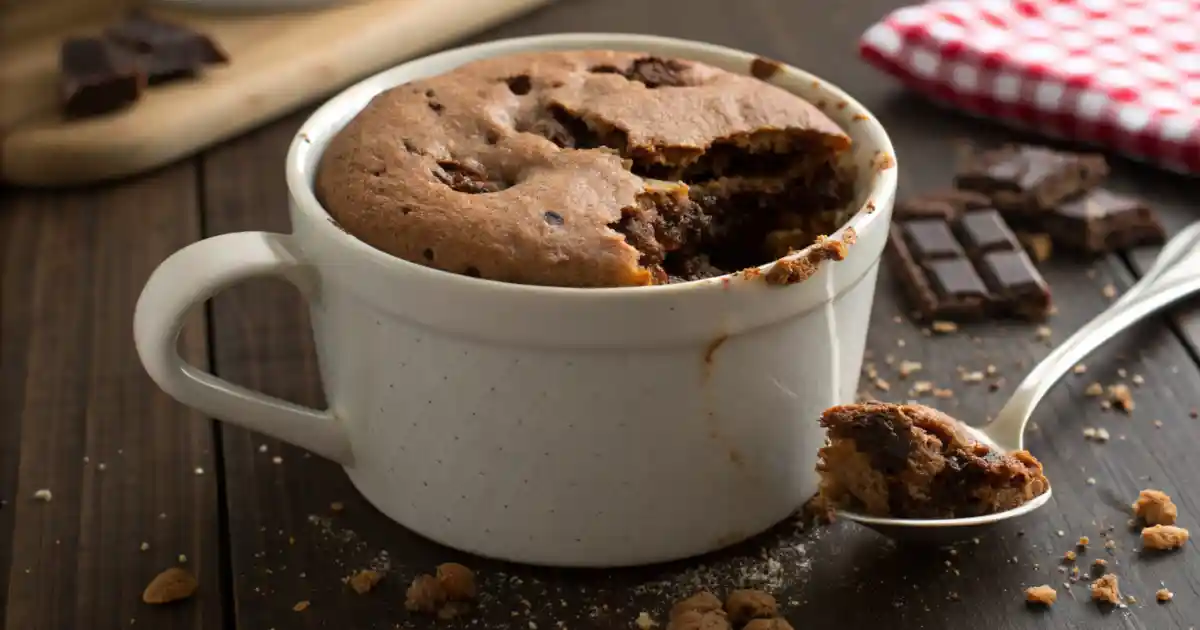 Freshly made protein mug cake no flour served in a ceramic mug, topped with chocolate chips, on a wooden kitchen table with a spoon nearby.