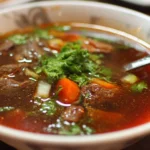 A bowl of hearty beef soup garnished with herbs and vegetables.